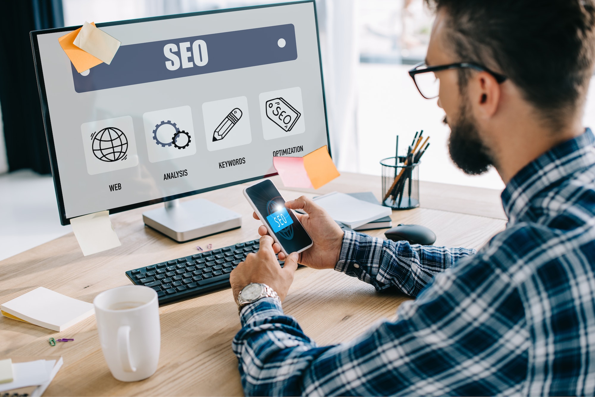 handsome young seo manager using smartphone and computer at workplace